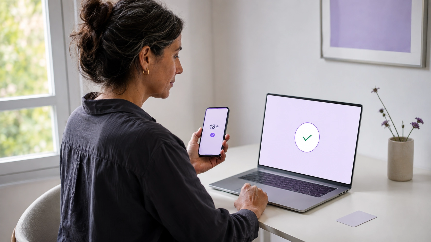 Someone holding a phone showing an 18+ confirmation while the laptop in front of them shows a green tick