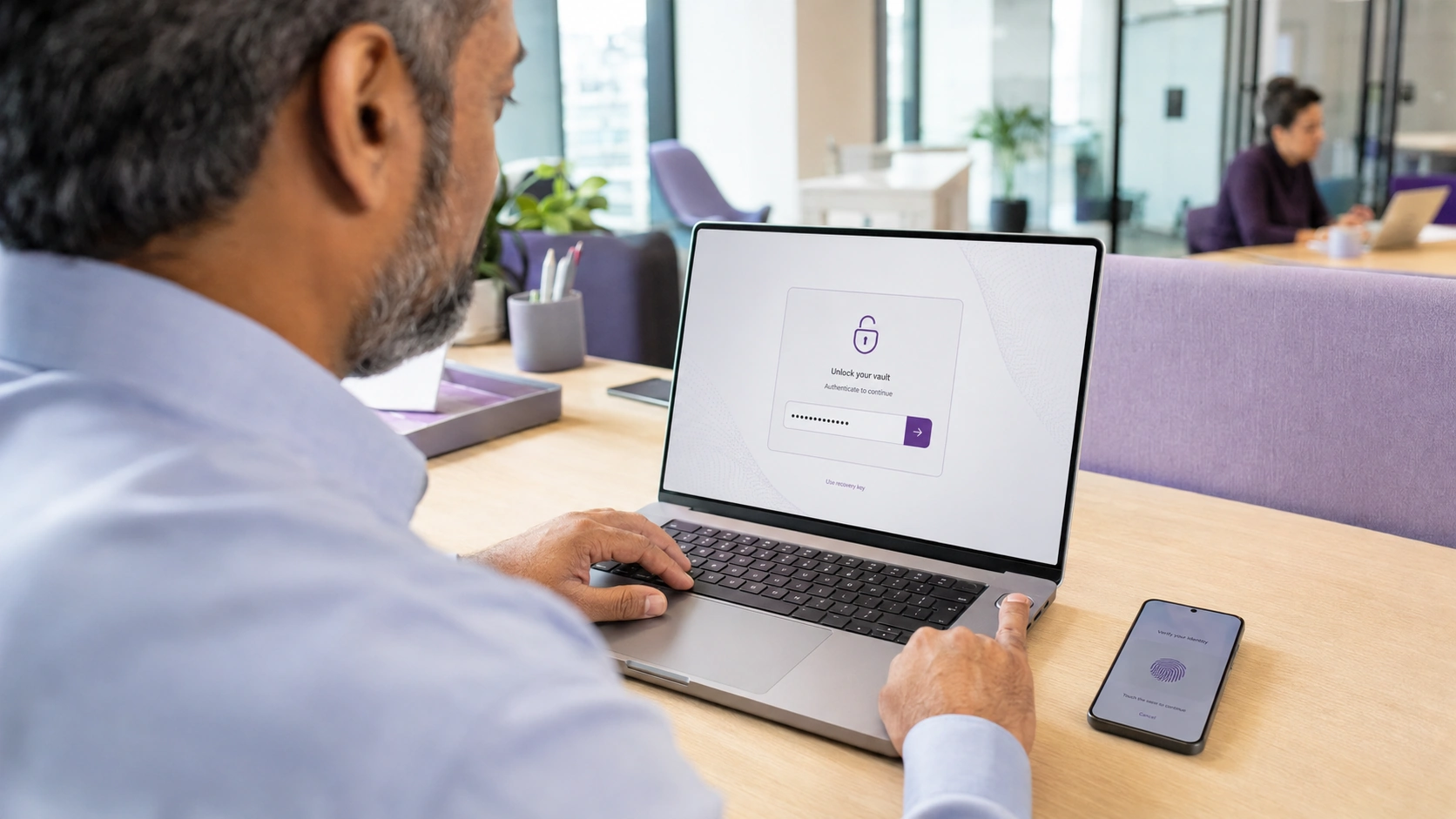 A person unlocking their password vault on a laptop, with a phone alongside asking them to confirm their identity
